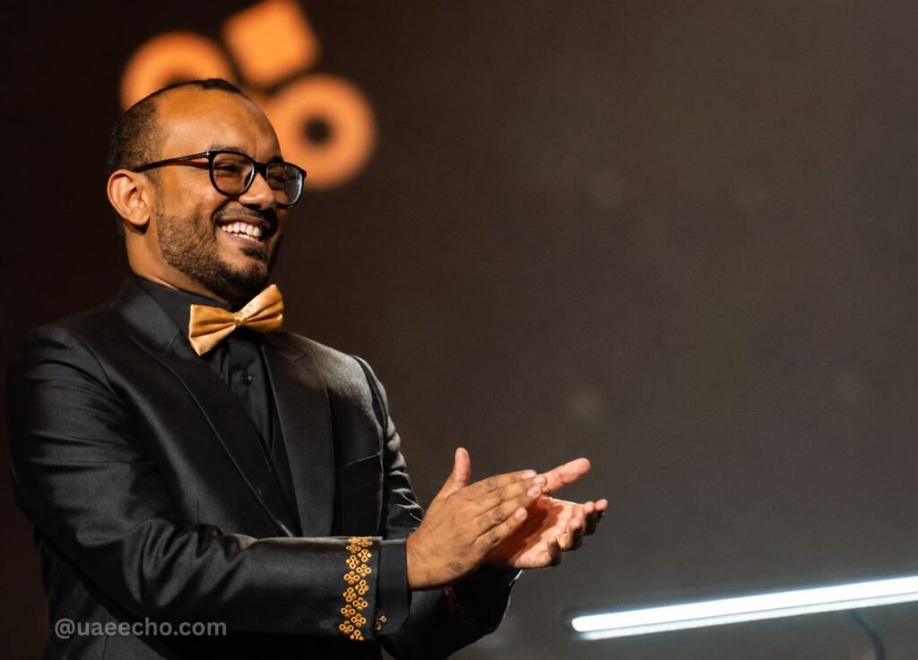 Smiling man in a black suit with a gold bow tie clapping on a stage with warm lighting.