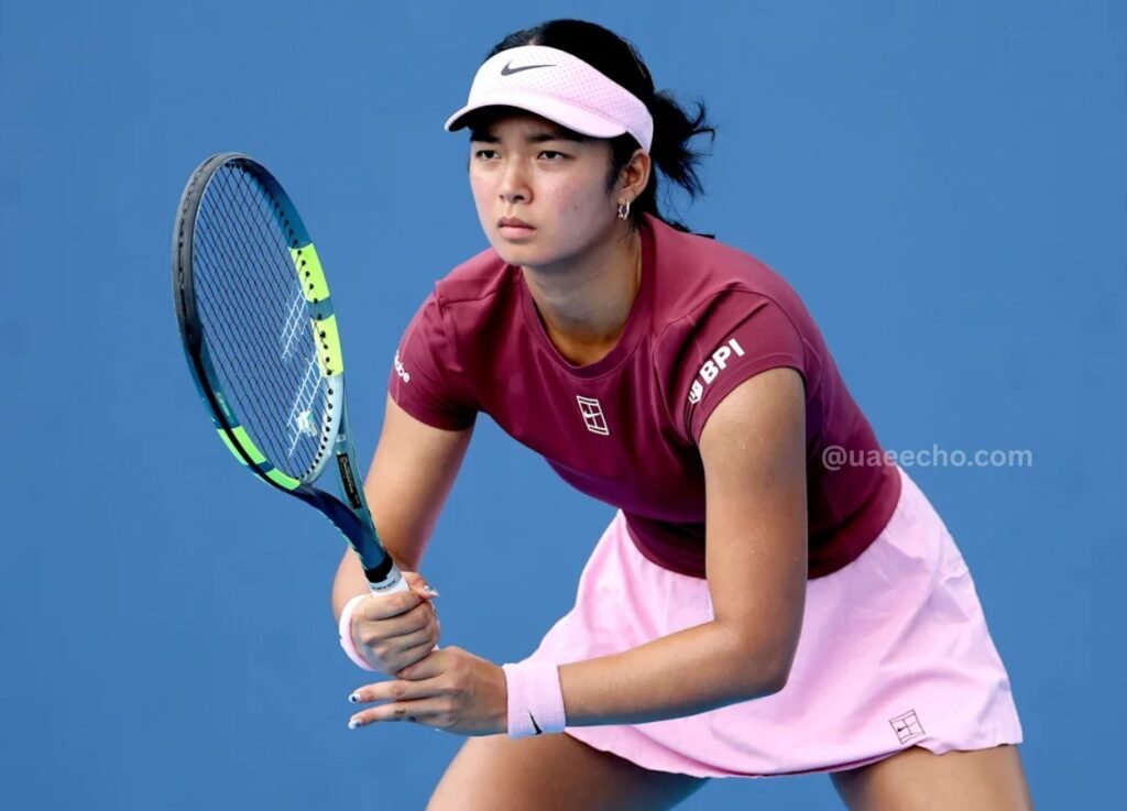 - UAE Echo Female tennis player in a pink visor, maroon shirt, and pink skirt, crouched and ready to return a ball with a blue racket on a blue court surface.